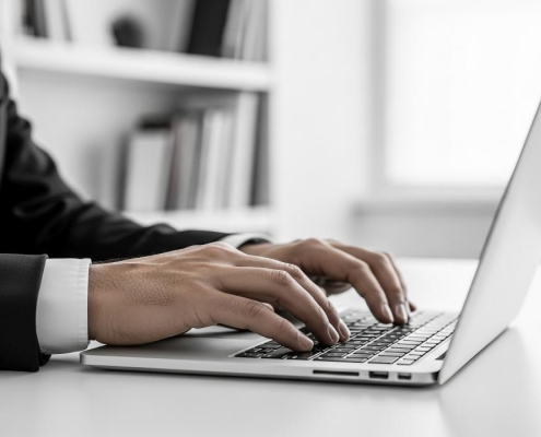Photo of close up of a mans hands typing on a laptop keyboard in an office setting with a bookshelf